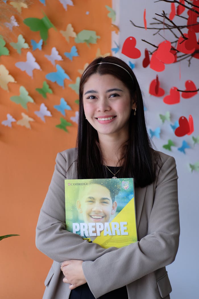 gallery-6 A young woman smiling while holding a Cambridge book in a classroom setting with colorful decor.